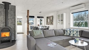 A modern living room with a stone fireplace, a grey sofa, a dining area, and a large window with a view of snow-covered trees. - 3 bedroom amazing home in Storvorde (Storvorde)