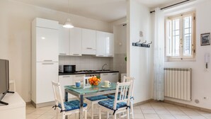 Une cuisine de taille réduite avec des meubles blancs, une table à manger en verre avec des coussins bleus, et un radiateur près d’une fenêtre.