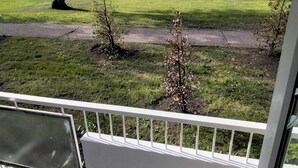 Vue depuis un balcon doté d’une balustrade blanche, donnant sur une cour verdoyante avec de petits arbres et une clôture en bois en arrière-plan.