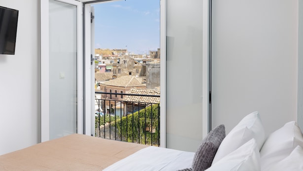 A bedroom with a bed, white pillows, and a view through an open sliding door to a balcony with a pool and buildings in the background. - City Panorama - One Bedroom Apartment (Corfu)