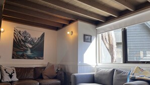 Una sala de estar con un sofá de cuero marrón, un sofá gris, una mesa de centro de madera, un cuadro de un paisaje montañoso y una ventana con cortinas.