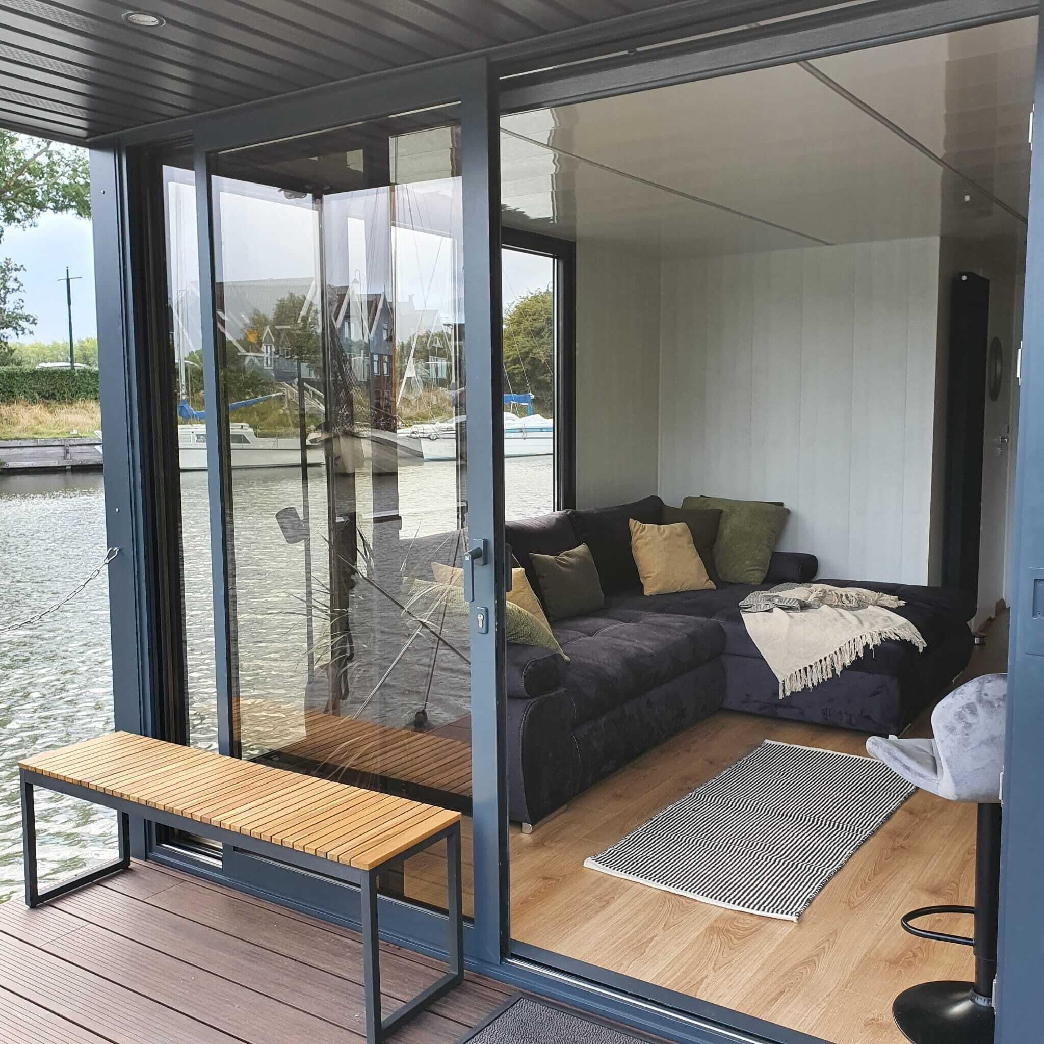 A modern living room with a dark-colored sofa, a wooden bench, and a glass door leading to a water body.