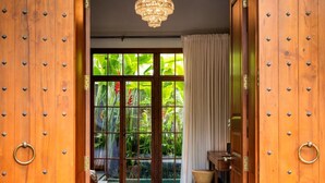 Una habitación con una puerta de madera grande, un candelabro, vista a exuberante vegetación a través de puertas de vidrio y una pequeña mesa de madera.