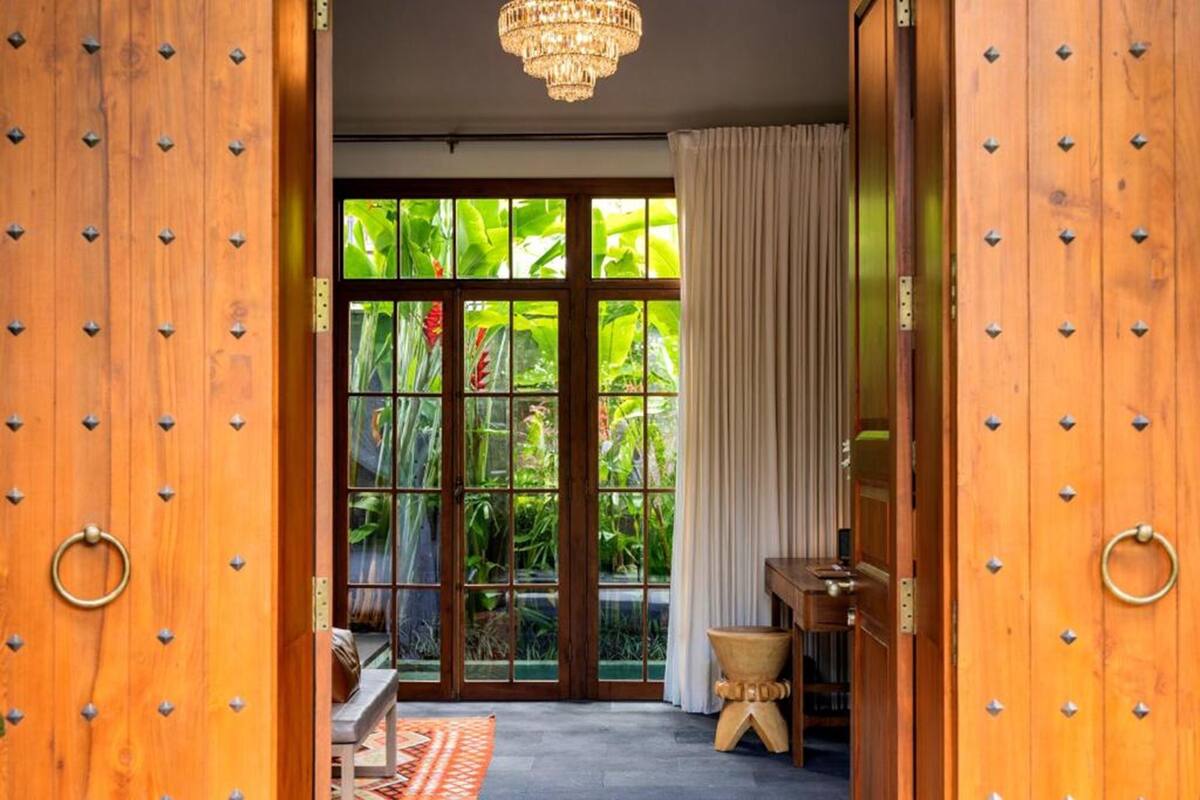 A room with a large wooden door, a chandelier, a view of lush greenery through glass doors, and a small wooden table.