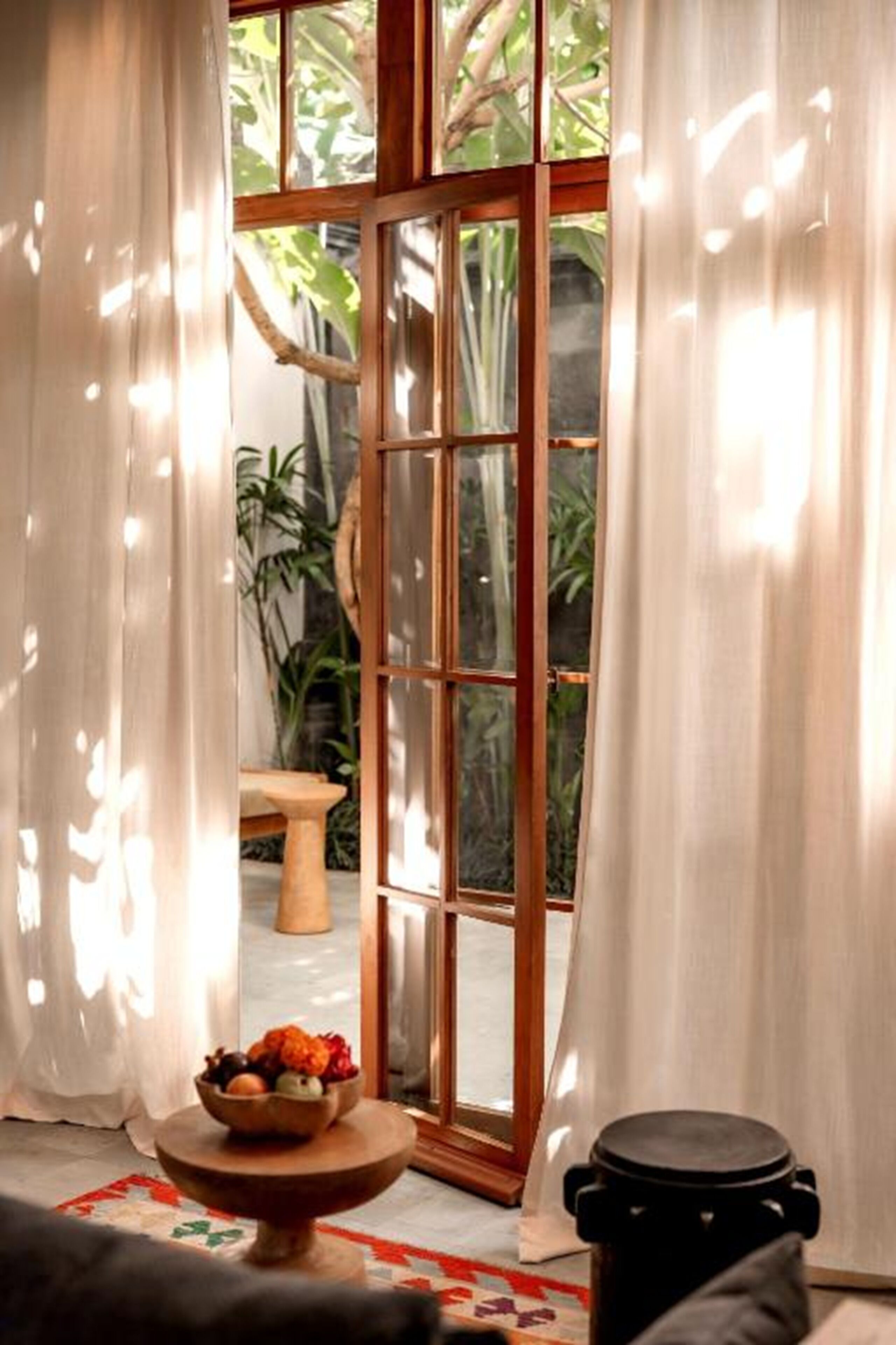 Une pièce dotée d’une grande porte vitrée donnant sur un jardin, une table ronde en bois sur laquelle se trouve un saladier rempli de fruits, et un récipient noir posé sur le sol.