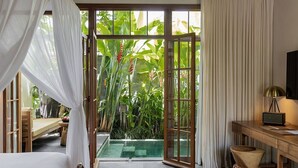 Una habitación con cama, escritorio, computadora, lámpara, un taburete pequeño, ventana con cortinas y vista a una piscina y vegetación.