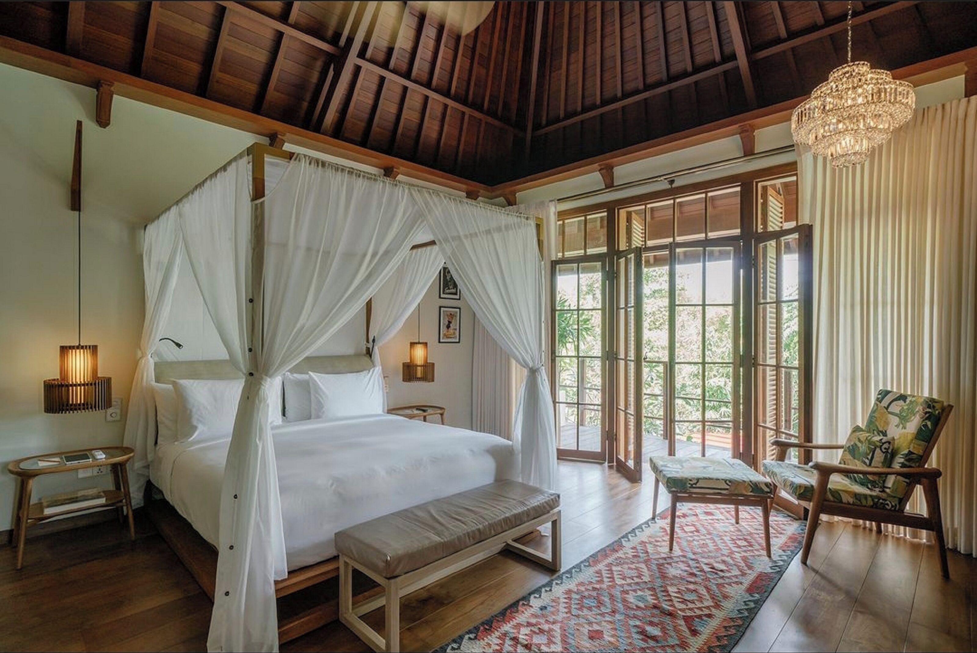 A bedroom with a four-poster bed, a bench, a chair, and a large window with curtains.