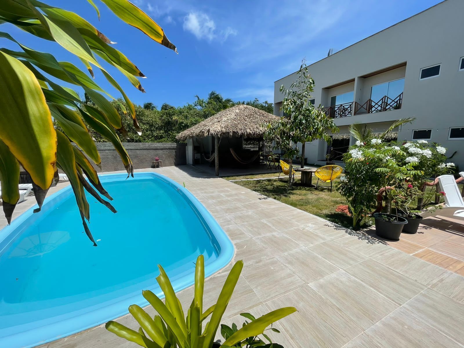 A swimming pool with a thatched-roof structure in the background, surrounded by a patio area with chairs and potted plants.