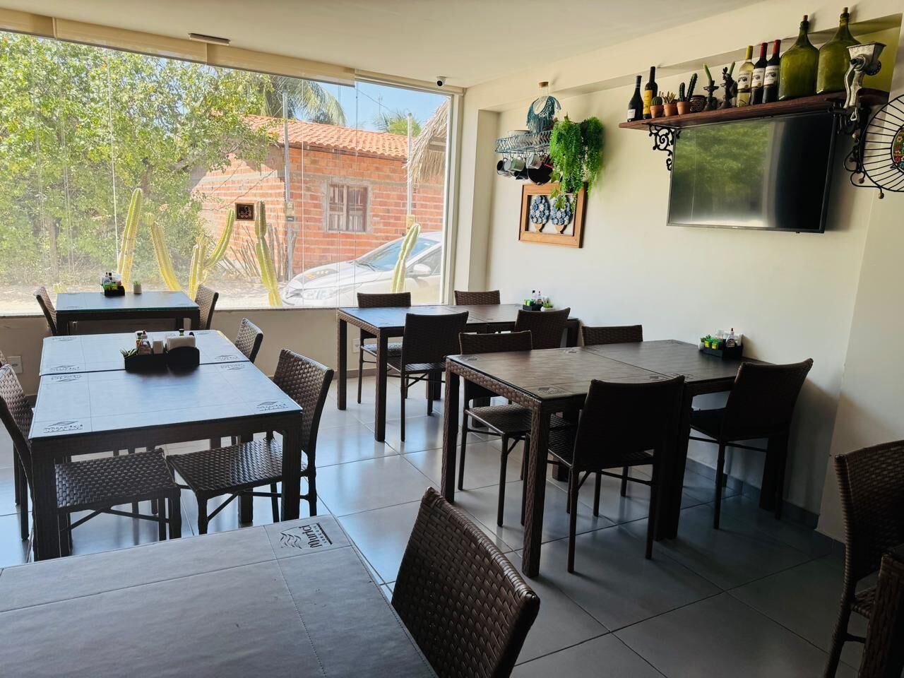A dining area with multiple tables and chairs, a television mounted on the wall, and a shelf with bottles and plants.