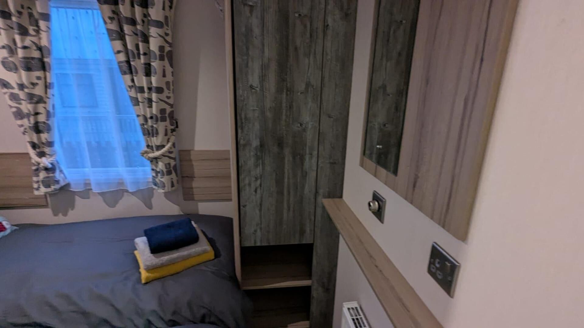 A small, narrow room with a bed, a wooden wardrobe, and a window with patterned curtains.
