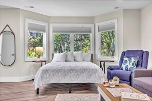 Une chambre avec un grand lit, deux fenêtres, une chaise, une table et un miroir.