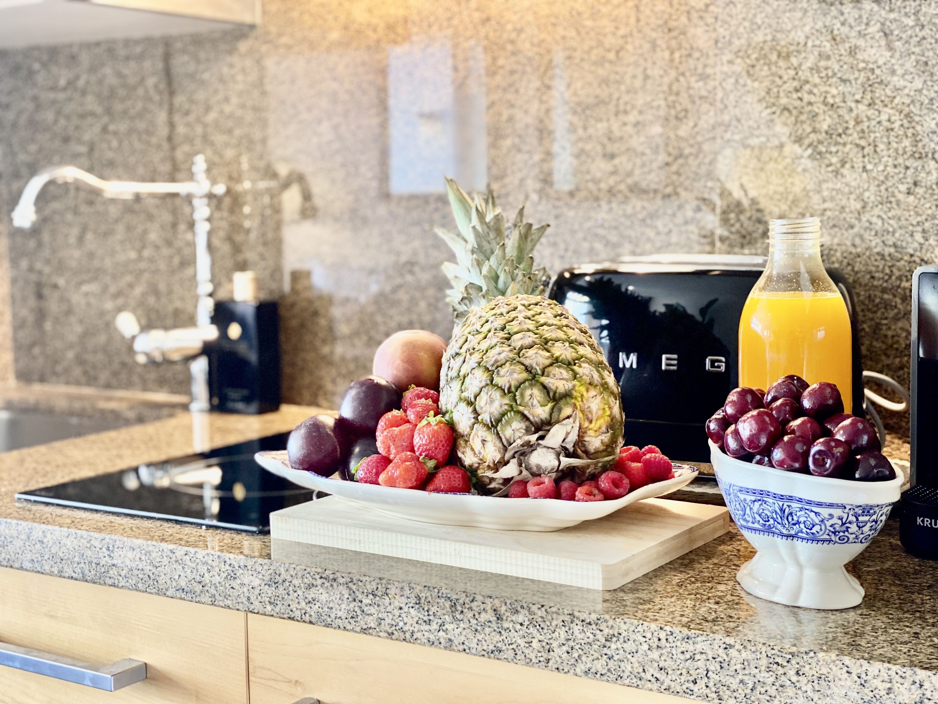 Una encimera de cocina con un frutero que contiene fresas, frambuesas y uvas, una piña y una botella de jugo de naranja.