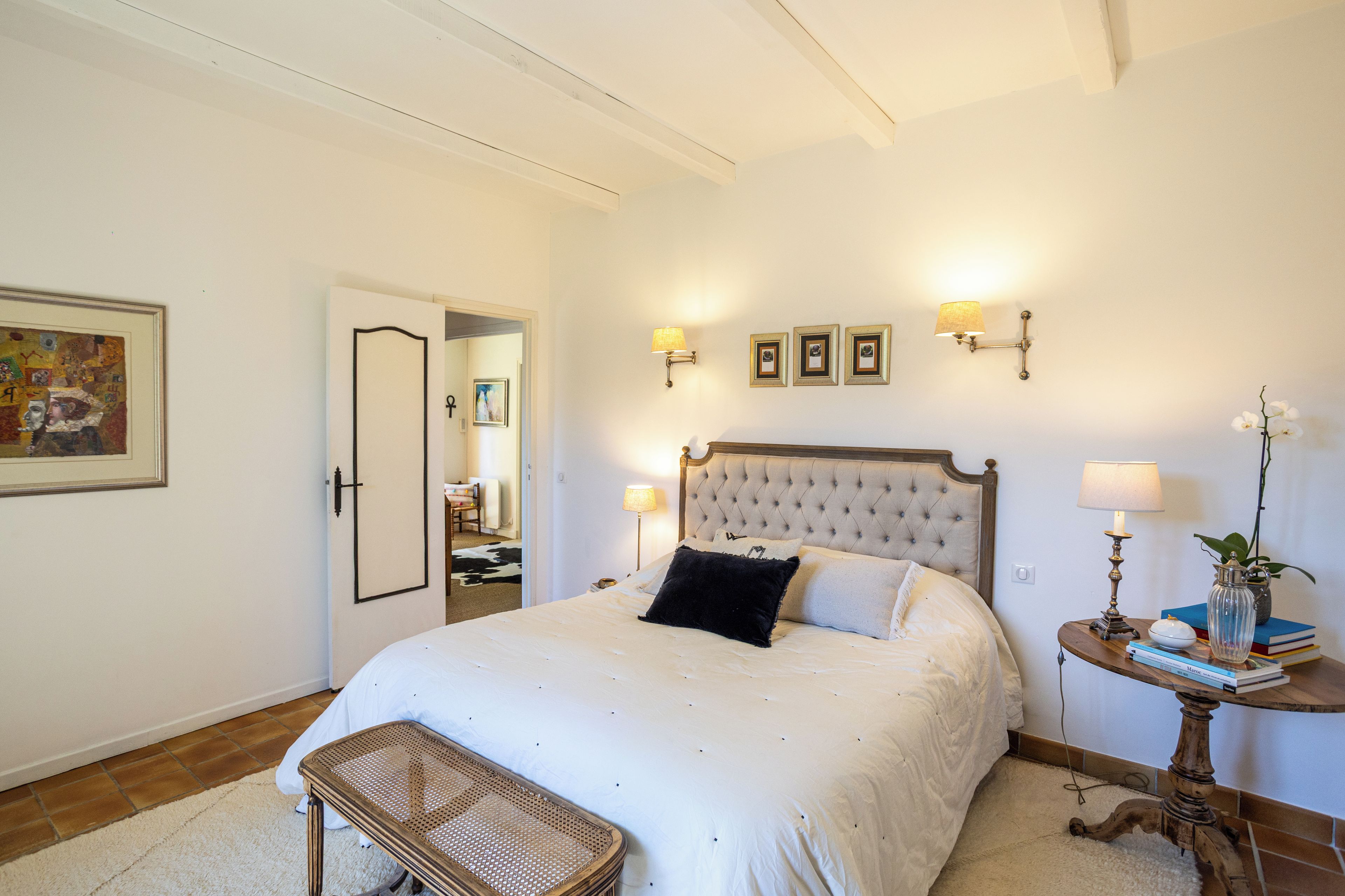 Une chambre à coucher avec un grand lit, des lampes de chevet, une petite table avec des livres et un vase, et une porte donnant sur une autre pièce.