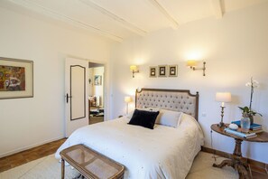 A bedroom with a large bed, bedside lamps, a small table with books and a vase, and a door leading to another room.