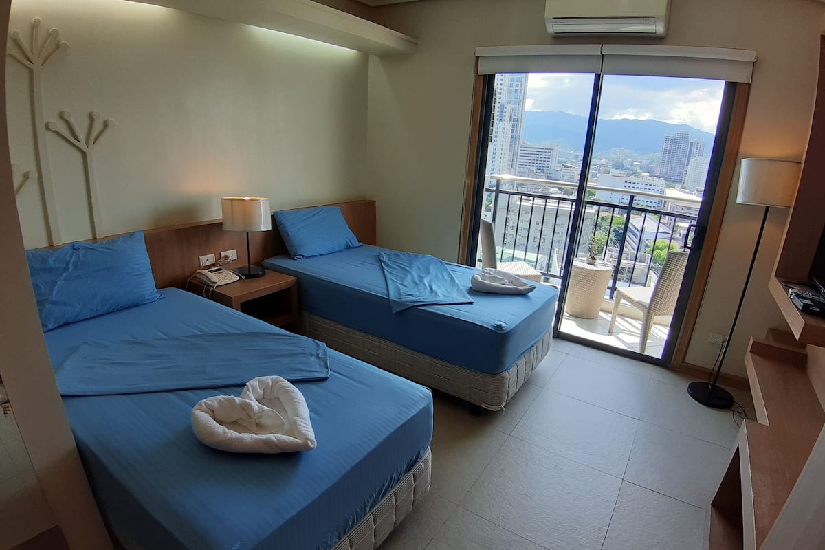 A hotel room with two beds, a balcony with a view of the city, and an air conditioning unit.