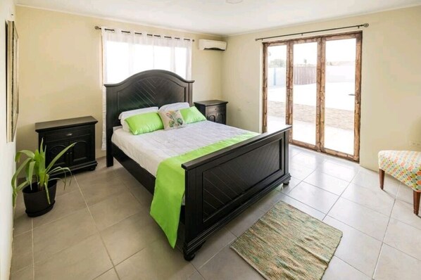 Un dormitorio con cama, mesitas de noche, una planta en maceta, un banco y un ventanal corredizo al exterior.