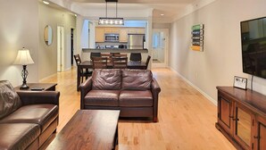 A modern living room with a leather sofa, wooden coffee table, and a TV on a wooden cabinet. A dining area with a kitchen in the background is visible.