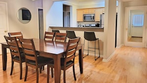A dining area with a wooden table and chairs, a kitchen in the background, and a hallway leading to a door.