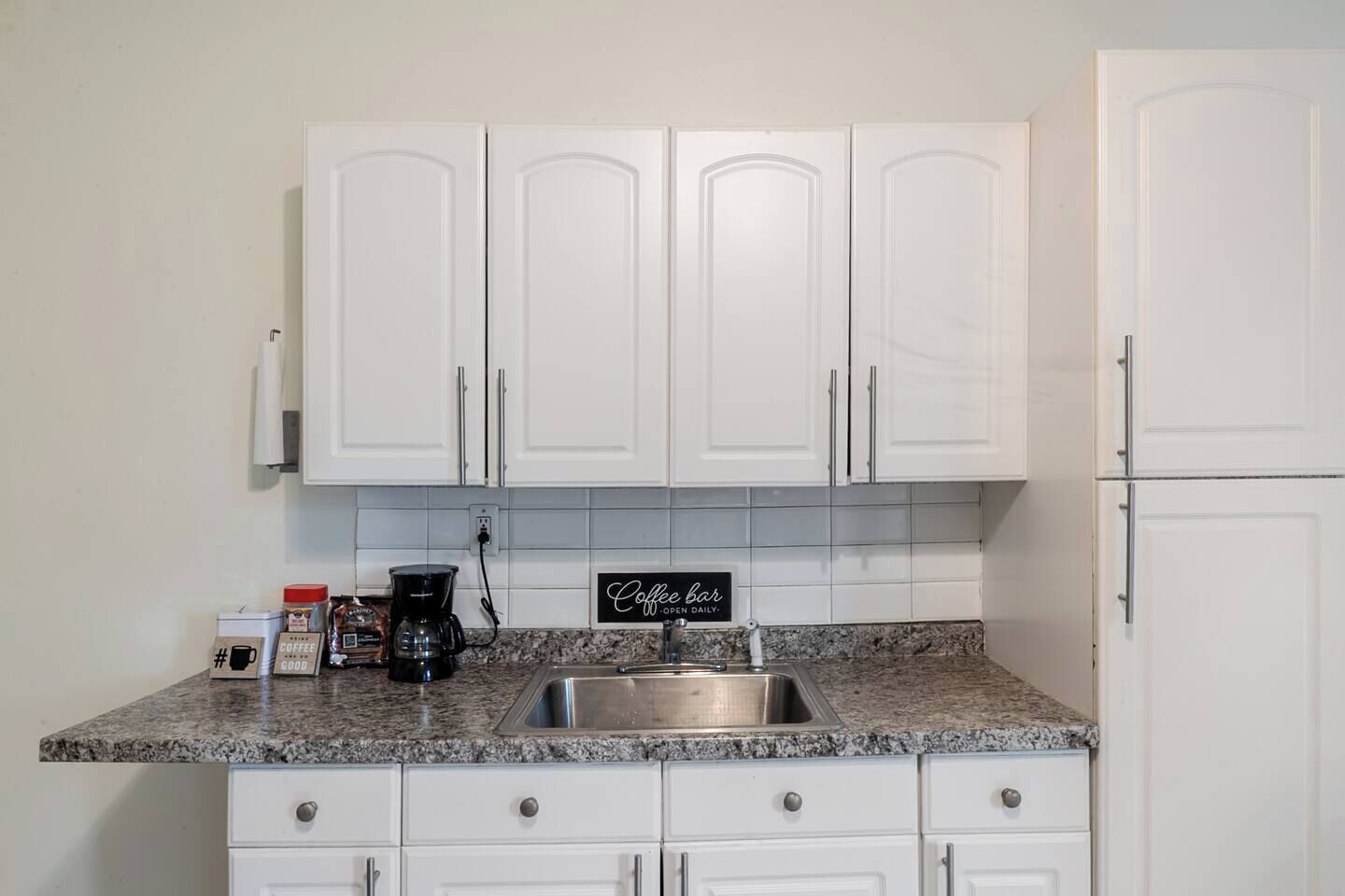 A kitchen with white cabinets, a granite countertop, a coffee maker, and a sign that reads 'Coffee bar open daily.'