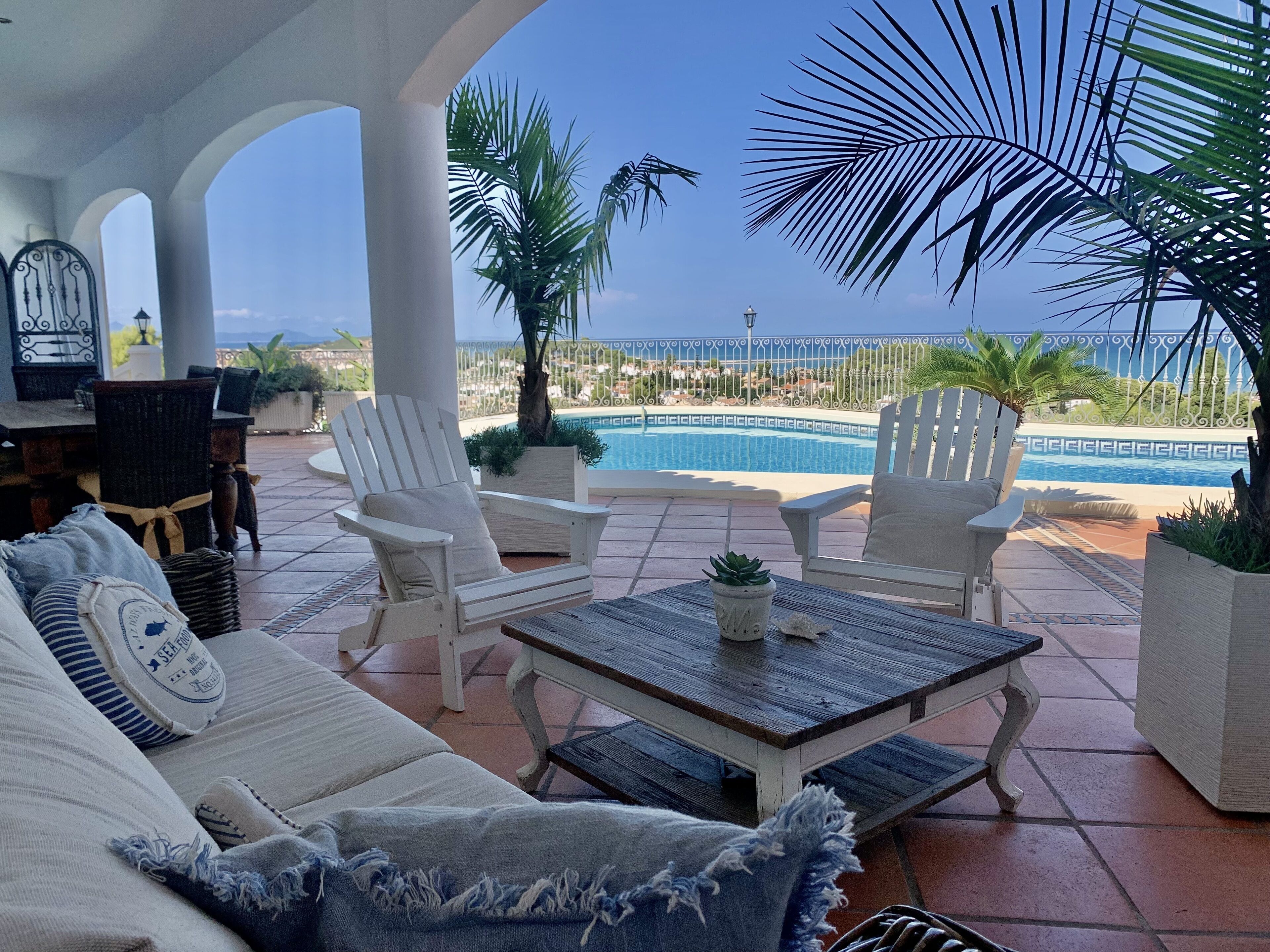 Ein überdachter Sitzbereich im Freien mit weißen Stühlen und einer Couch, ein Pool und Blick auf den Ozean.