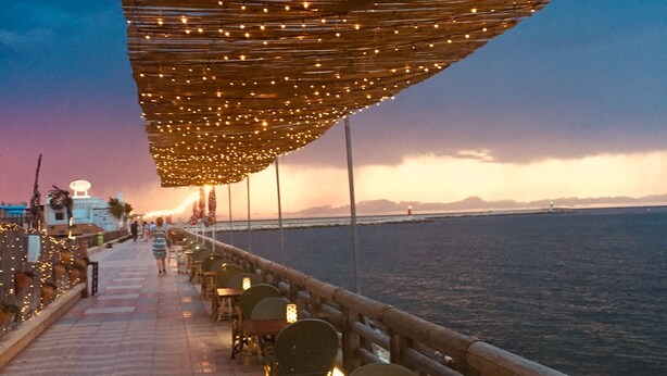 A seaside restaurant with outdoor seating, a view of the sea, and a wooden pergola with string lights. - 400 m2 dream villa / 180 degree panoramic sea view, in top location!  (Dénia)