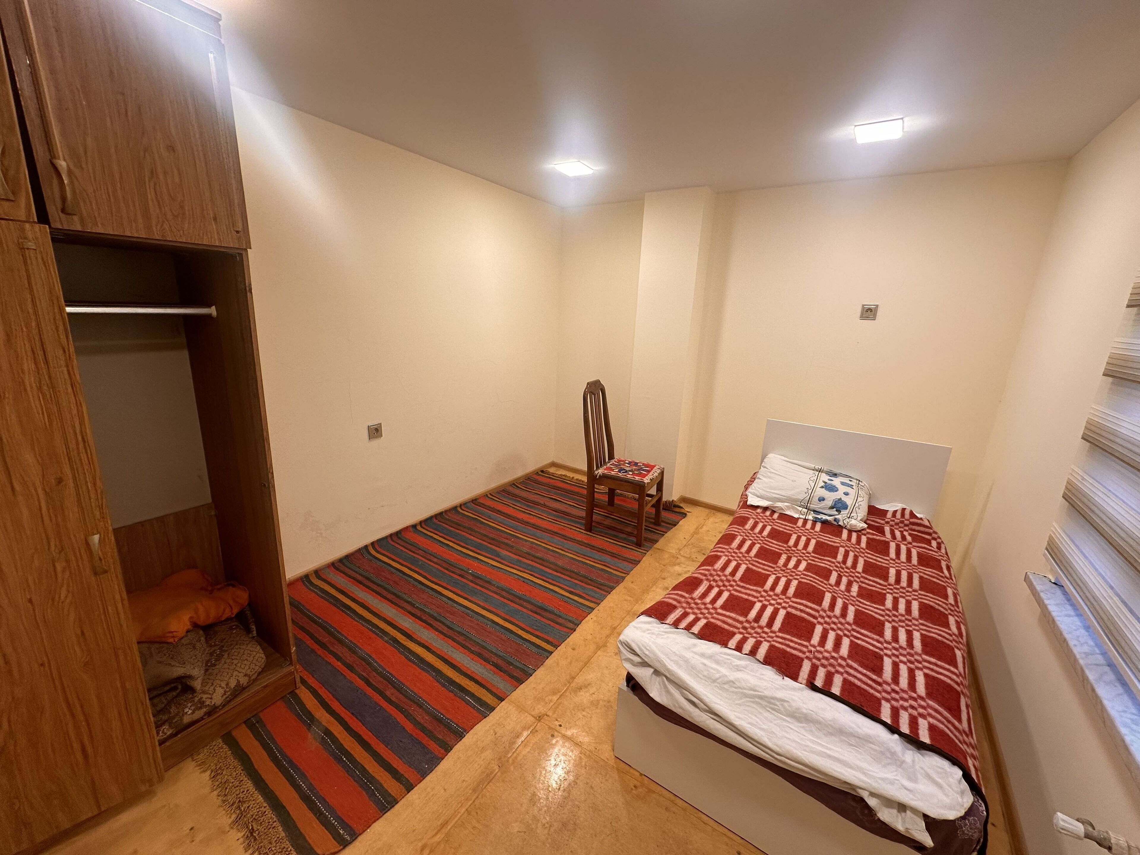 Een kleine, goed verlichte kamer met een bed, een stoel, een houten kledingkast en een gestreept tapijt.