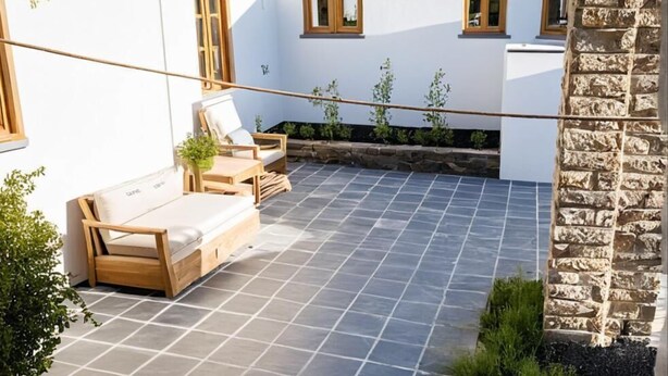 Eine überdachte Terrasse mit gefliestem Boden, Holzmöbeln und einer Steinwand.