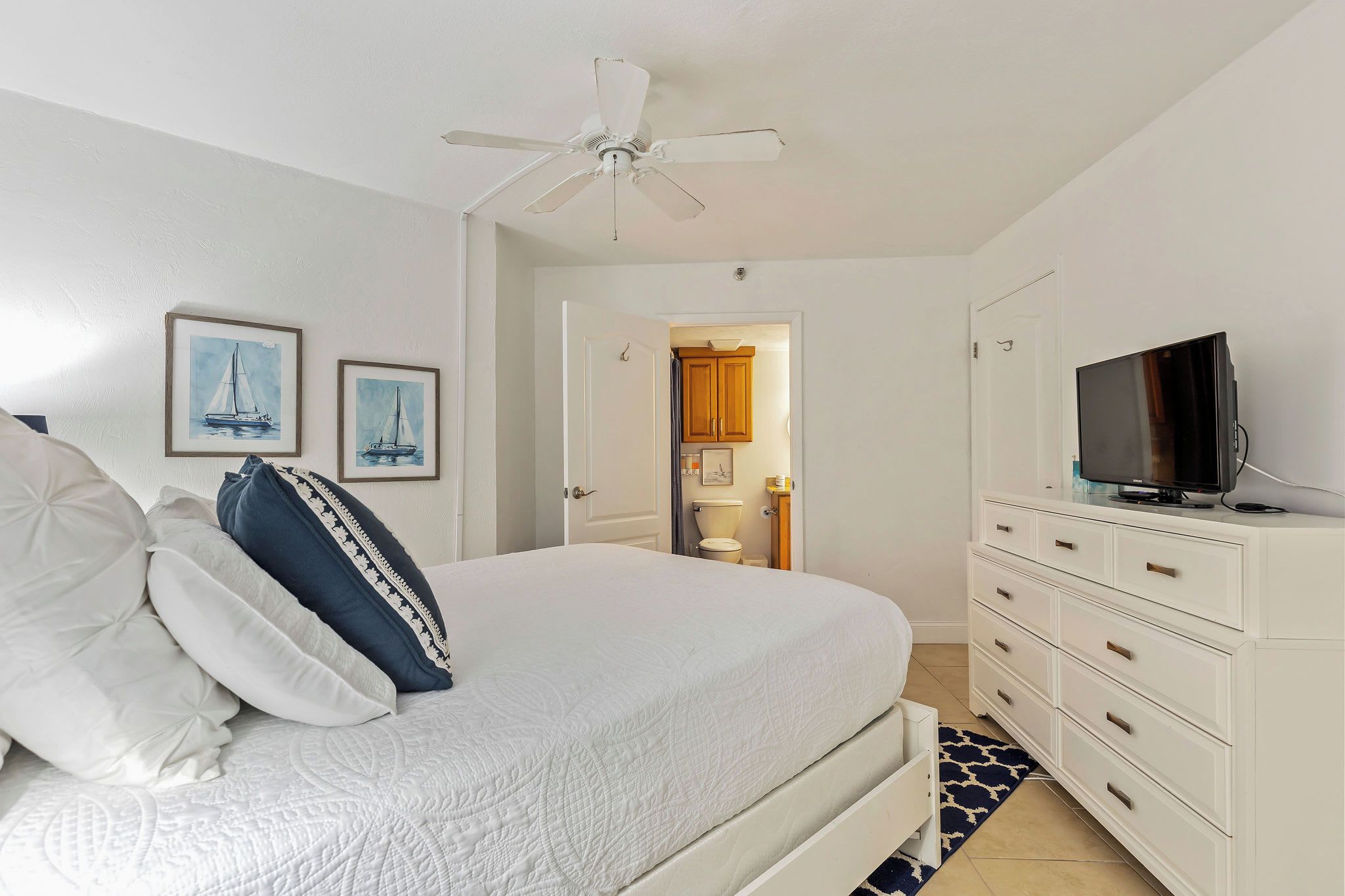A bedroom with a bed, a ceiling fan, a TV on a dresser, and framed artwork on the wall.