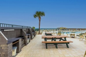 An outdoor patio with barbecue grills, picnic tables, and lounge chairs by a pool. - Ocean View Suite w/ Pool, Hot Tub & Beach Access (Daytona Beach Shores)