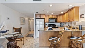 A kitchen with wooden cabinets, a central island with bar stools, a refrigerator, a microwave, and a dining area with a table and chairs. - Ocean View Suite w/ Pool, Hot Tub & Beach Access (Daytona Beach Shores)