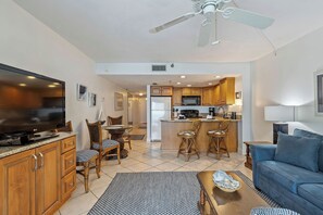 A modern living room with a TV, wooden cabinets, a blue sofa, and a kitchen area with a microwave and refrigerator. - Ocean View Suite w/ Pool, Hot Tub & Beach Access (Daytona Beach Shores)