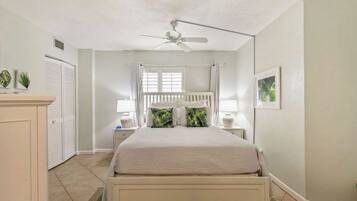 Une chambre à coucher comprenant un lit, un ventilateur de plafond, une fenêtre avec des rideaux, une table de chevet et une commode.