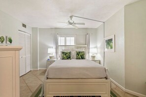 Une chambre à coucher comprenant un lit, un ventilateur de plafond, une fenêtre avec des rideaux, une table de chevet et une commode.