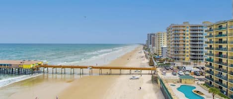 Un hotel frente a la playa con un muelle que se adentra en el océano, un área de piscina y varios balcones.