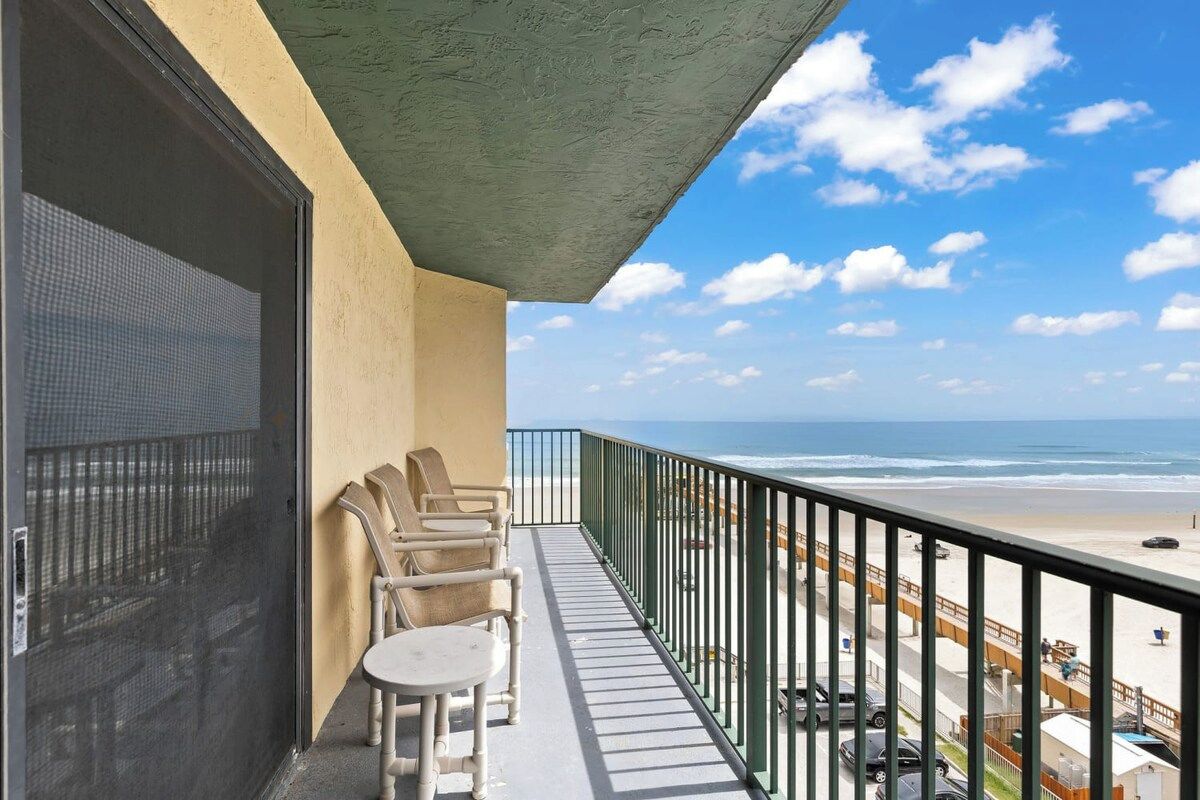 A balcony with a view of the beach, a chair, and a small table.