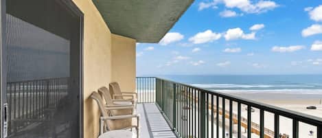 A balcony with a view of the beach, a chair, and a small table.