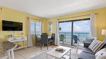 Ein Wohnzimmer mit Fernseher, Esstisch, Stühlen, einer Couch und einem Balkon mit Meerblick.