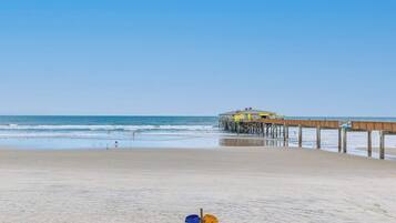 A beach with a wooden pier extending into the ocean, a building on the pier, and a blue and yellow bucket on the sand.