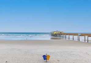 A beach with a wooden pier extending into the ocean, a building on the pier, and a blue and yellow bucket on the sand. - Breathtaking Views, Heated Pool & Beach Access! (Daytona Beach Shores)
