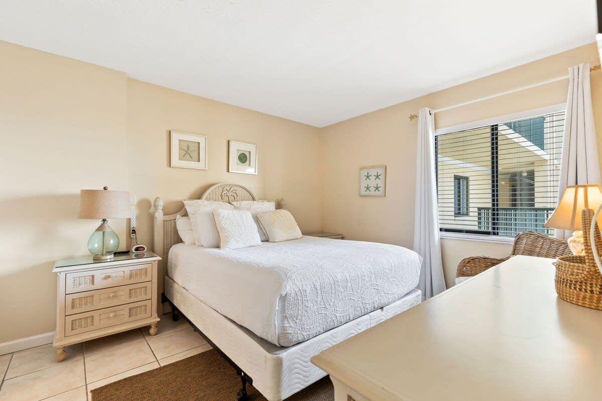 Quarto com cama, mesa de cabeceira, luminária e janela com cortinas.