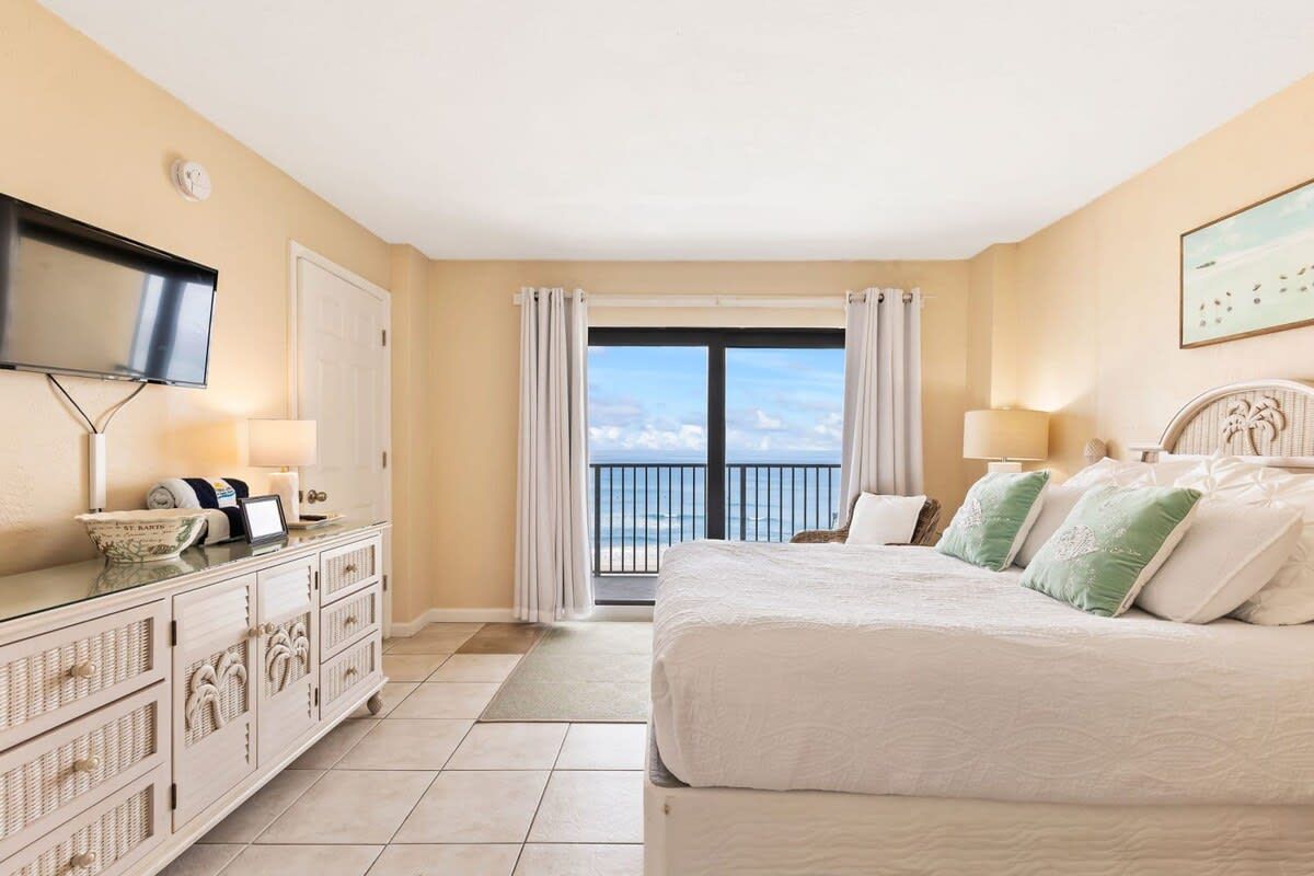 Quarto de hotel com uma cama grande, televisão presa na parede, cômoda e varanda com vista para o mar.