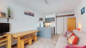 A compact living space with a wooden dining table, a sofa with colorful cushions, a television, a kitchen area, and a ceiling light fixture.