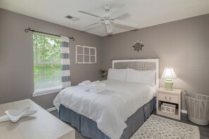 Ein Schlafzimmer mit einem Bett, Nachttischen, einer Lampe, einem Deckenventilator, einem Fenster mit Jalousien und einer Wanddekoration mit Schiffslenkrad.