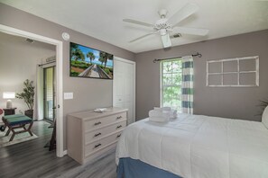 Un dormitorio con cama, cómoda, ventilador de techo, ventana con cortinas y puerta que da a otra habitación.