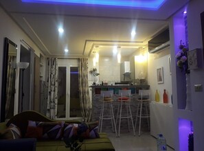 A modern living room with a sofa, a dining area with bar stools, a kitchen with a bar counter, and a sliding glass door leading to a balcony.