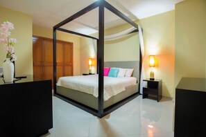 A four-poster bed with a white bedspread and colorful pillows, flanked by bedside tables with lamps, in a room with wooden furniture and a vase of flowers.