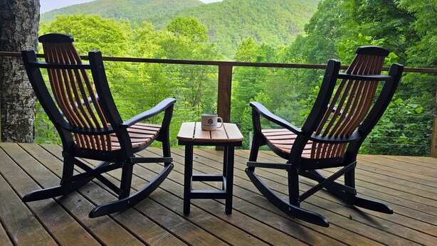 Two rocking chairs and a small table on a wooden deck with a view of a forested mountain range.