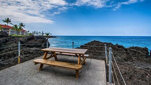 A wooden picnic table on a concrete platform with a view of the ocean and a building with a red roof in the background.