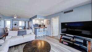 A modern living room with a sofa, coffee table, TV, and kitchen area in the background. - Breathtaking Ocean View, Beachfront Condo-Steps to Beach/Pools, Sea Colony East (Bethany Beach)