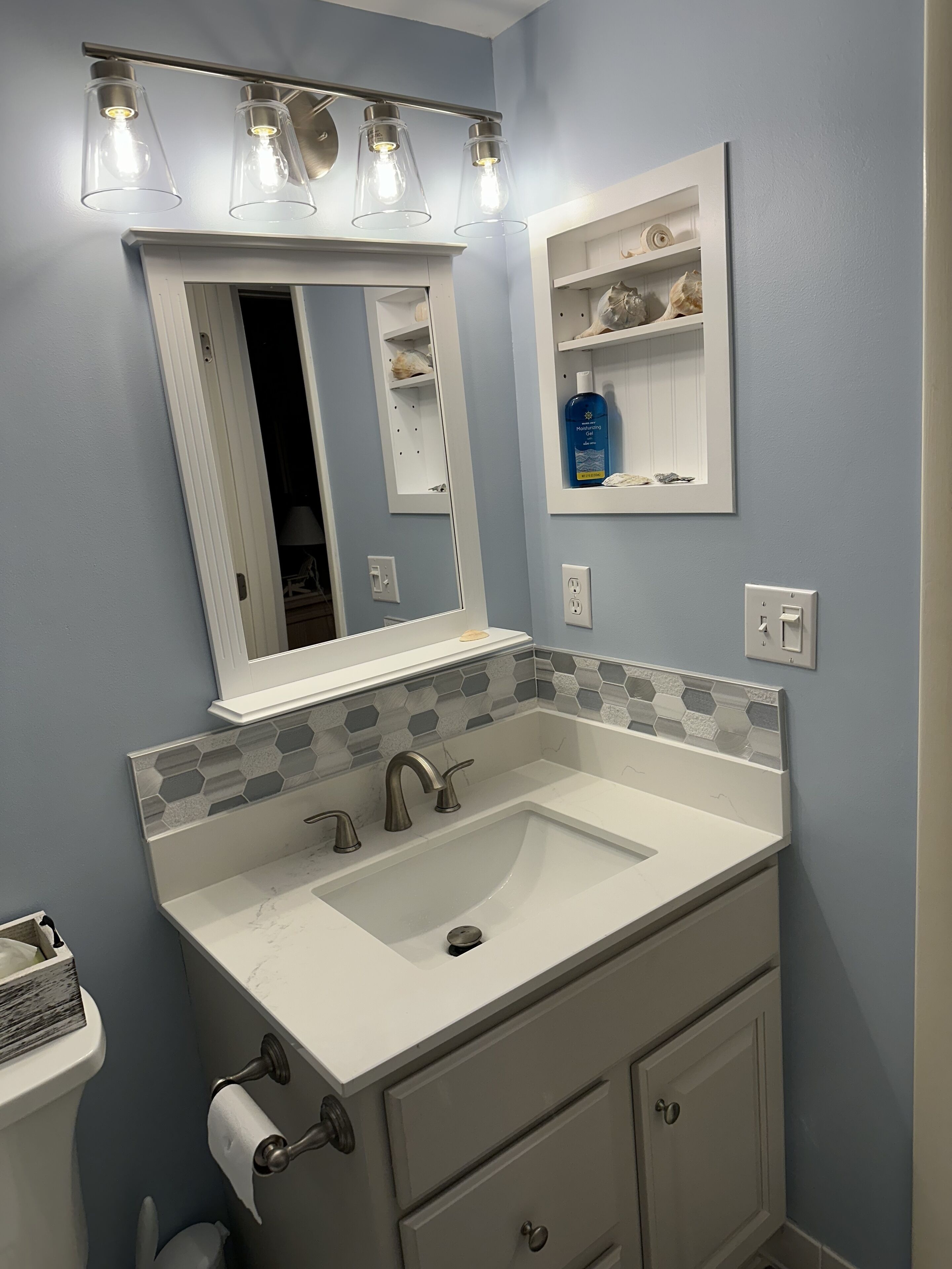 Un baño con un lavabo blanco, dos grifos, un espejo con marco blanco y un estante con artículos de aseo.